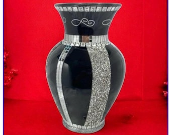 Puede incluir: Un jarrón decorativo negro con detalles de mosaico plateado. El jarrón tiene una abertura ancha, un cuello estrecho y un cuerpo redondeado. Rayas de mosaico plateado y una banda de mosaico rodean el jarrón. Fondo rojo.