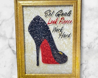 May include: A framed glitter art piece with a black high heel shoe with a red sole. The text "Set Goals Look Fierce Work Hard" is written in black and red glitter.