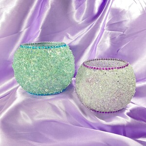 May include: Two decorative glass candle holders, one is teal and the other is purple with iridescent glitter. Both have a rhinestone trim, the teal one has a teal rhinestone trim and the purple one has a purple rhinestone trim.