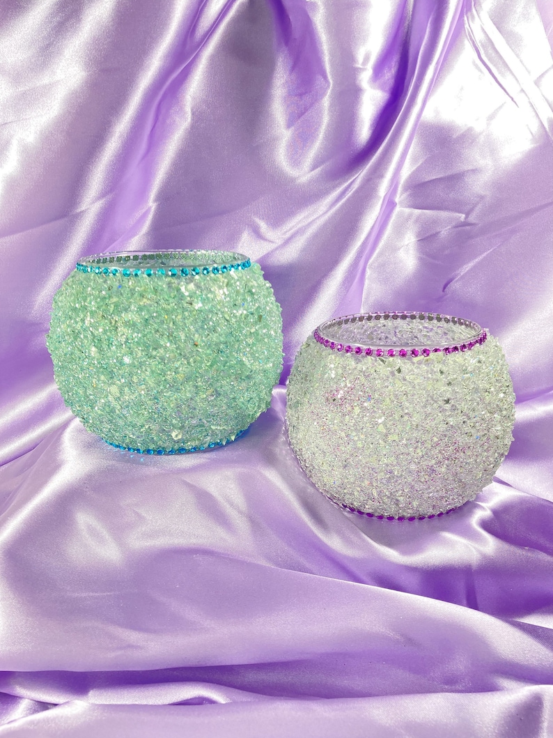 May include: Two decorative glass candle holders, one is teal and the other is purple with iridescent glitter. Both have a rhinestone trim, the teal one has a teal rhinestone trim and the purple one has a purple rhinestone trim.