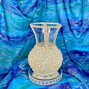 May include: A clear glass vase with a rounded, textured body covered in small, sparkling embellishments. The rim and a vertical line down the front are adorned with rhinestones. The vase sits on a mirrored base with a decorative, jeweled border.