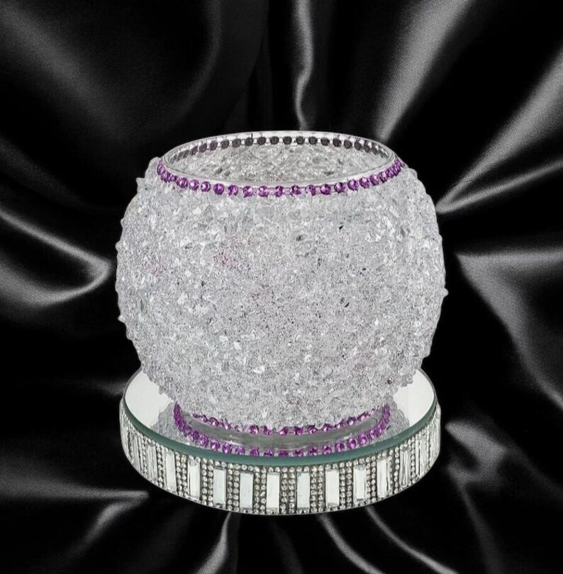May include: A clear glass candle holder with a purple rhinestone trim. The candle holder is sitting on a mirrored base with a silver rhinestone trim