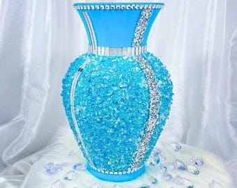 Puede incluir: Un jarrón decorativo azul con una superficie texturizada similar a un cristal. Acentos plateados y pedrería adornan el borde y los lados. El jarrón está sobre una superficie blanca y esponjosa, con cristales dispersos.