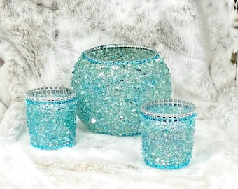 May include: A set of three glass candle holders in varying sizes, featuring a textured, crushed light blue glass finish and a silver-toned metallic rim adorned with small, clear, reflective circular accents around the top edge.