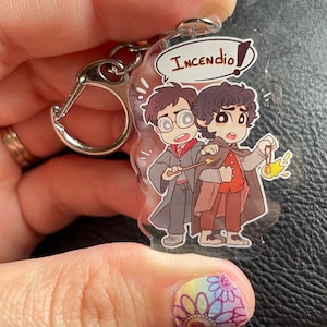May include: A clear acrylic keychain featuring two cartoon characters, one with brown hair and glasses, the other with brown hair, holding a wand and a small flame. The text "Incendio!" is in a speech bubble above the characters.