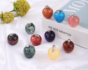 May include: A collection of gemstone apple pendants in various colors, including red, green, yellow, and blue. Each apple has a silver-colored bail. The pendants are displayed on a white surface with a flower and a book in the background.