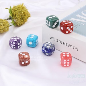 May include: A collection of colorful gemstone dice. The dice are in various colors, including green, red, purple, blue, and pink, with white dots. The dice are arranged on a white surface. The text "WE GTE NEWTON" is visible.