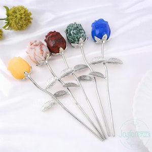 May include: Five gemstone rose hair sticks in yellow, pink, red, green, and blue. Each rose has a silver-colored stem and leaves. The roses are arranged on a white surface.