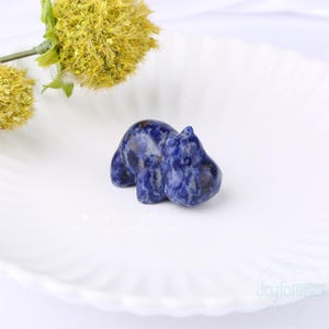 May include: A small, carved hippo figurine made of blue sodalite stone. The hippo is in a resting pose, with its legs tucked under its body. The stone has a marbled appearance with white and dark blue swirls. The figurine is on a white, ridged plate.