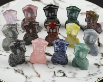 May include: A collection of 14 small, carved figurines made from various types of polished stones. The figurines are all in the shape of a human torso and are arranged in a circle on a white marble surface.