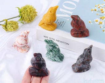May include: A collection of carved stone bird figurines in various colors, including yellow, red, green, and marble. The figurines are displayed on a white surface with a book and flowers in the background. The text "MORE" and "WE GTE NEWTON" are visible.