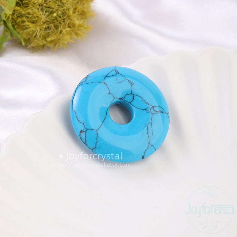 Puede incluir: Una piedra turquesa en forma de rosquilla con vetas oscuras. La piedra tiene un acabado pulido y brillante y un agujero en el centro. La piedra est&aacute; sobre un plato blanco festoneado. El texto de la imagen dice "Joyforcrystal" y una direcci&oacute;n web.
