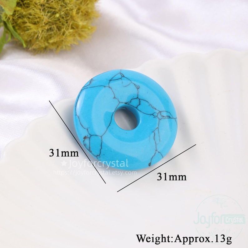 Puede incluir: Una gema turquesa en forma de rosquilla con vetas negras. La piedra mide 31 mm de di&aacute;metro y pesa aproximadamente 13 g. La imagen incluye el texto "Joyforcrystal" y una direcci&oacute;n web.