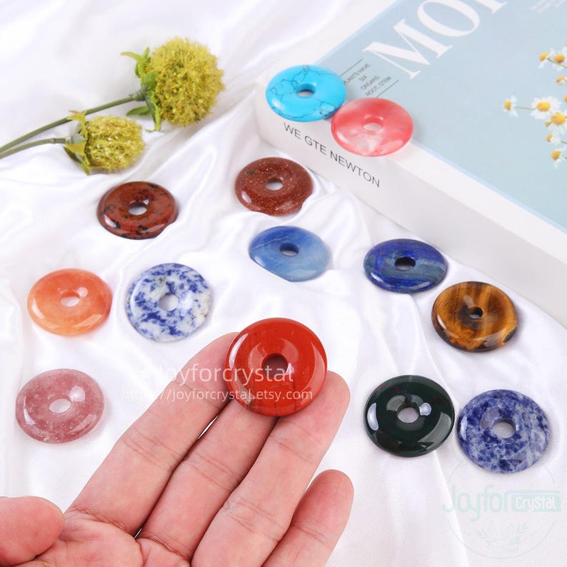 Puede incluir: Una colecci&oacute;n de colgantes de piedras preciosas en forma de rosquilla de colores, en varios colores, incluyendo rojo, azul y marr&oacute;n. Algunos colgantes se sostienen en una mano. Los colgantes se exhiben sobre una superficie blanca con un libro en el fondo. El texto "Joyforcrystal" es visible.