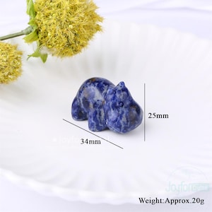 May include: A small, blue and white sodalite hippo figurine. The hippo is 34mm long and 25mm tall, with a weight of approximately 20g. The figurine is set on a white, scalloped plate, with a yellow flower in the background.