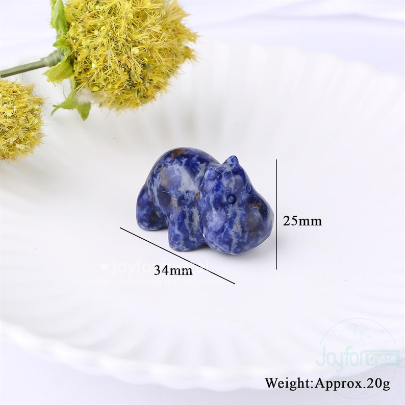 May include: A small, blue and white sodalite hippo figurine. The hippo is 34mm long and 25mm tall, with a weight of approximately 20g. The figurine is set on a white, scalloped plate, with a yellow flower in the background.