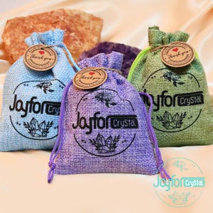 May include: Three drawstring bags in blue, purple, and green, each with a circular logo that reads "Joyfor Crystal." The bags have a textured appearance and are adorned with a small tag that says "Thank You." The bags are tied with matching colored drawstrings.