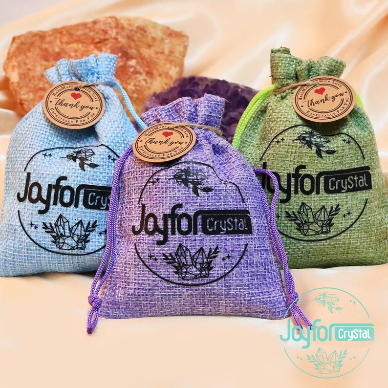 May include: Three drawstring bags in blue, purple, and green, each with a circular logo that reads "Joyfor Crystal." The bags have a textured appearance and are adorned with a small tag that says "Thank You." The bags are tied with matching colored drawstrings.