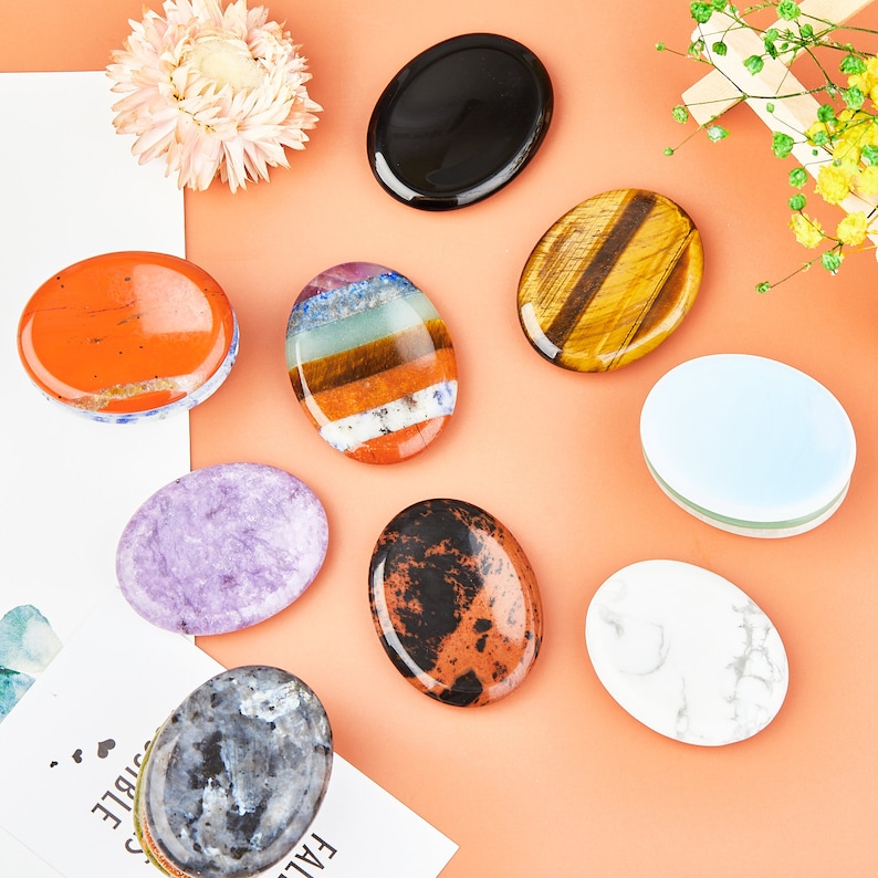 May include: Assortment of eight oval-shaped polished stones, each with a different color and pattern. The stones are arranged on a peach-colored surface.