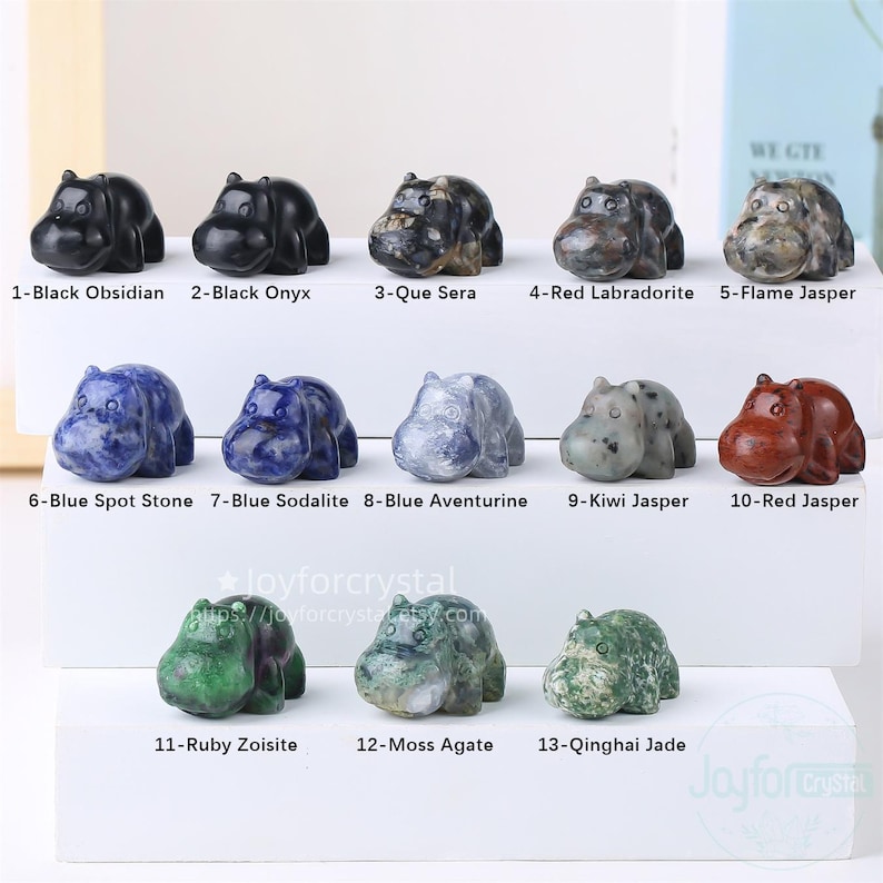 May include: A collection of thirteen gemstone hippo figurines in various colors and patterns. The figurines are labeled with the type of stone, including Black Obsidian, Red Jasper, and Blue Spot Stone. The figurines are displayed on a white surface.
