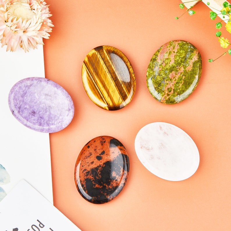 May include: Five oval-shaped polished stones in various colors and patterns. The stones are arranged on a light orange surface. The stones are: purple, tiger's eye, green, white, and red with black.