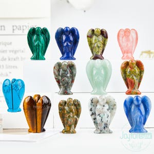 Puede incluir: Una colección de pequeñas figuras de ángeles talladas en varias piedras, mostrando una variedad de colores. Los ángeles tienen alas y túnicas detalladas, con tonos de verde, azul, rosa y marrón. Las figuras se exhiben sobre una superficie blanca.