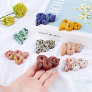May include: A selection of carved stone snake figurines in diverse colours, such as blue, green, yellow, red, and speckled patterns. The figurines are presented on a white surface with a book and flowers in the background. The text "WE GTE NEWTON" is visible.