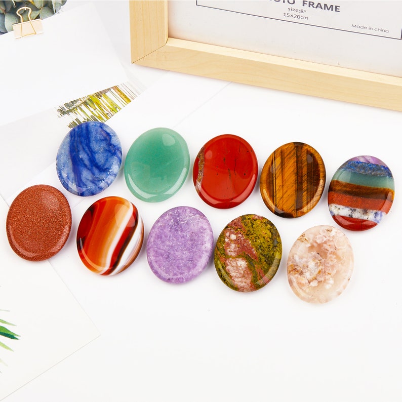 May include: Ten smooth, oval-shaped polished stones in various colors, including red, green, blue, purple, brown, and white. The stones are arranged in a row on a white surface.