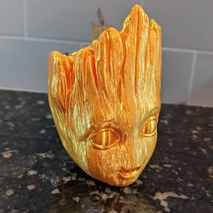May include: A gold-colored Groot-shaped planter with a textured surface. The planter has a smiling face and holds a small plant. The planter is made of a shiny material and is sitting on a dark surface.