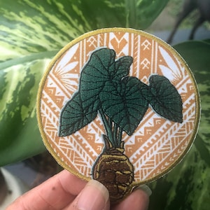 May include: A round, embroidered patch with a green plant with brown roots on a white and orange background. The patch has a gold border.