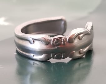 May include: A silver ring made from a bent spoon with a decorative design. The ring is open ended and adjustable.