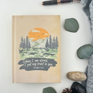 May include: A brown journal featuring a landscape scene of mountains, trees, and a river. The text "When I am afraid, I put my trust in you. -Psalm 56:3" is printed on the front cover.