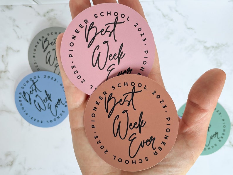 K&ouml;nnte beinhalten: Vier runde Aufkleber mit dem Text "Pioneer School 2023" und "Best Week Ever" in einer handschriftlichen Schriftart. Die Aufkleber sind in rosa, blau, braun und gr&uuml;n.