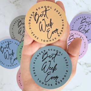 K&ouml;nnte beinhalten: Eine Hand h&auml;lt f&uuml;nf runde Aufkleber mit dem Text "Pioneer School Best Week Ever" in schwarzer Schrift. Die Aufkleber sind in verschiedenen Farben: hellblau, hellgr&uuml;n, helllila, hellorange und hellrosa.