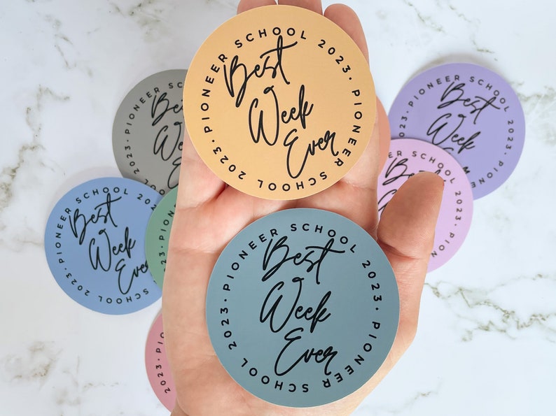 K&ouml;nnte beinhalten: Eine Hand h&auml;lt f&uuml;nf runde Aufkleber mit dem Text "Pioneer School Best Week Ever" in schwarzer Schrift. Die Aufkleber sind in verschiedenen Farben: hellblau, hellgr&uuml;n, helllila, hellorange und hellrosa.