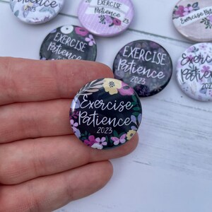 May include: A set of six round buttons with a floral design and the text "Exercise Patience 2023". The buttons are black with white lettering and have a variety of colorful flowers.