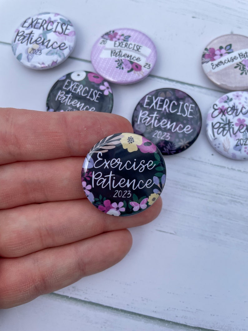 May include: A set of six round buttons with a floral design and the text "Exercise Patience 2023". The buttons are black with white lettering and have a variety of colorful flowers.