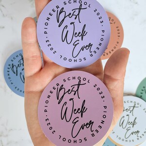 K&ouml;nnte beinhalten: Drei runde Aufkleber mit dem Text "Best Week Ever" in einem Kreis mit den Worten "Pioneer School 2023" au&szlig;en herum. Die Aufkleber sind in Lila, Rosa und Gr&uuml;n.