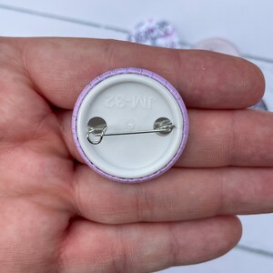 May include: The back of a purple button with a white center. The button has a safety pin attached and the text "JM-82" printed on the center.