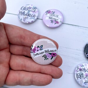 May include: Three round buttons with a floral design and the text "Exercise Patience" in white. The buttons are on a white wooden surface.