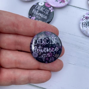 May include: A black and purple button with a floral design and the text "EXERCISE patience 2023".