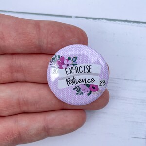 May include: A purple button with a white chevron pattern and floral accents. The button says "Exercise Patience" with the years 20 and 23 on either side of the text.