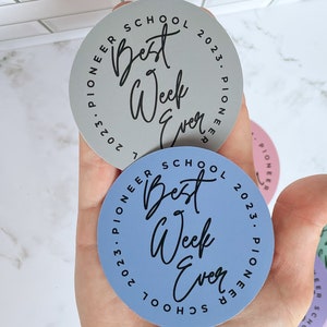 K&ouml;nnte beinhalten: Vier runde Aufkleber mit dem Text "Best Week Ever" in schwarzer Schrift auf blauem, grauem, rosa und gr&uuml;nem Hintergrund. Der Text "2023. Pioneer School" ist in Schwarz um jeden Aufkleber herum geschrieben.