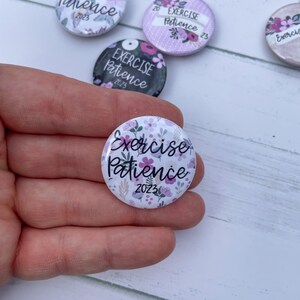 May include: Four white round buttons with floral designs and the text "Exercise Patience 2023". One button is held in a hand.