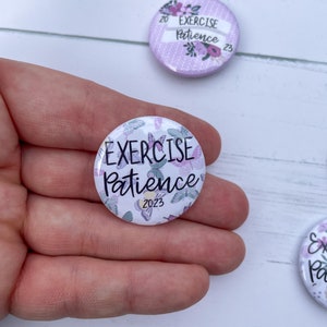 May include: A white button with a floral design and the text "Exercise Patience 2023" in black lettering.