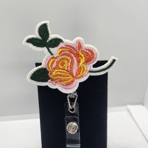 Könnte beinhalten: Ein weiß gesticktes Blumenabzeichen mit einer rosa und orangefarbenen Rose und grünen Blättern, befestigt an einem durchsichtigen Plastik-Badgehalter mit einem silbernen Clip.