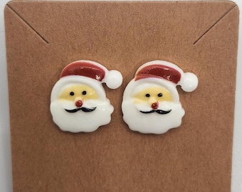 May include: A pair of white Santa Claus face stud earrings with red hats and yellow faces. The earrings are on a brown background.