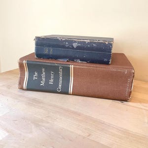 Może przedstawiać: Dwie stare książki ułożone na drewnianej powierzchni. Dolna książka jest brązowa ze złotymi literami: "The Matthew Henry Commentary". Górna książka jest mniejsza, ciemnoniebieska, z wyblakłym tekstem.