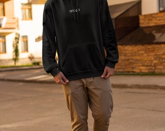 May include: A black hooded sweatshirt with a white inscription that reads "90:4:1". The model is wearing the sweatshirt with brown pants and white sneakers.