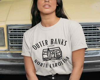 May include: A woman wearing a gray baseball cap and a white t-shirt with the text "Outer Banks Pogue Life North Carolina" and a black and white illustration of a van with a surfboard on top.